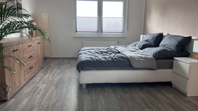 Een moderne slaapkamer met een groot bed en donkere beddengoed. Een commode en een plant completeren de inrichting.