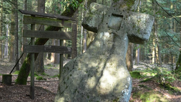Großer, moosbedeckter Steinkreuz im Wald neben einem hölzernen Wegweiser.