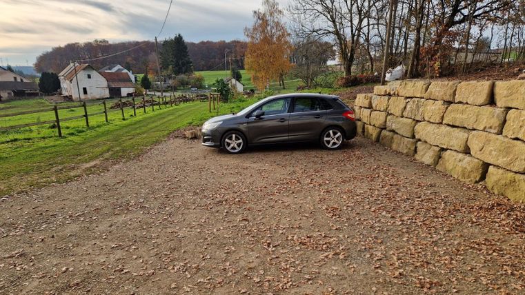 Een auto staat op een grindplaats in een landelijke omgeving. Op de achtergrond zijn weilanden en enkele gebouwen te zien.