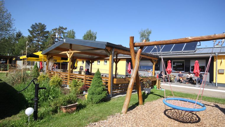 Ein einladendes Restaurant mit einer großen Terrasse und sonnigem Wetter. Im Vordergrund befindet sich eine Schaukel und grüne Bäume.