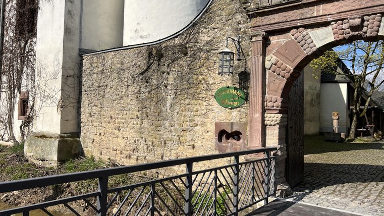 Un bâtiment historique avec une belle façade en pierre et une porte romantique. Un pont traverse un petit ruisseau menant à l'entrée.