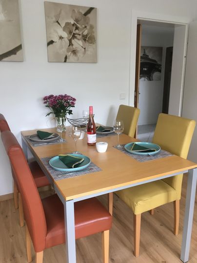 Une table à manger dressée avec des chaises colorées et un vase avec des fleurs. Sur la table se trouvent des verres et une bouteille de vin rosé.