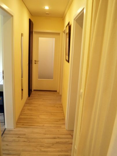 Un couloir lumineux avec des murs jaunes et un sol en bois moderne. Sur le côté gauche, il y a plusieurs portes.