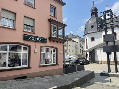 Eine Strasse mit einem griechischen Restaurant namens "Zorbas" und einer Kirche im Hintergrund. Der Himmel ist blau und es sind einige Autos auf der Straße.