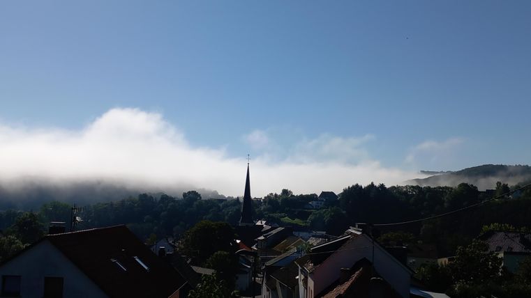A picturesque view with houses in the foreground and a church tower. In the background, dense fog is visible over the hills and a blue sky.