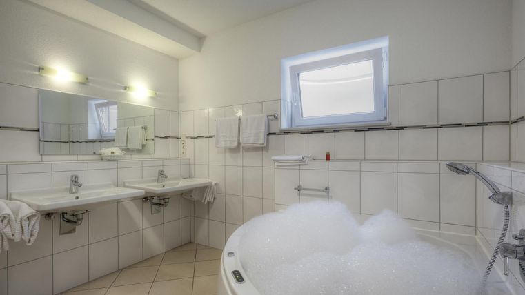 Ein modernes Badezimmer mit einer Badewanne voller Schaum. Das Zimmer hat helle Fliesen und einen großen Spiegel über den Waschbecken.