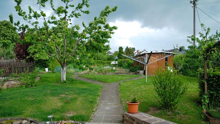 Ein ruhiger Garten mit einem Pfad, der durch das Grün führt. Im Hintergrund sind Bäume und ein Wäscheplatz zu sehen.