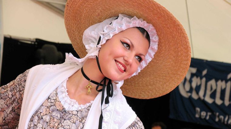 Eine Frau in traditioneller Tracht mit einem großen Strohhut lächelt fröhlich. Im Hintergrund sind unscharfe Menschen und eine dekorative Bühne sichtbar.