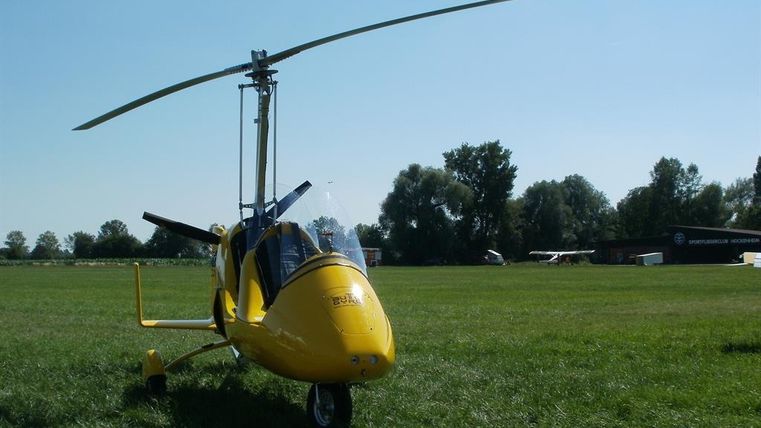 Ein gelbes Gyrocopter auf einer Wiese. Im Hintergrund sind Bäume und ein weiterer Luftfahrzeug zu sehen.