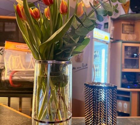 Een glazen vaas met kleurrijke tulpen en eucalyptus staat op een tafel. Ernaast staat een elegante kandelaar.