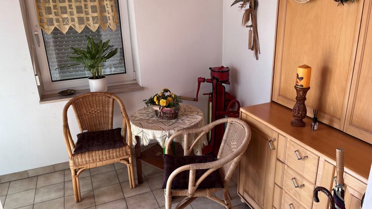 Eine gemütliche Sitzecke mit zwei Rattanstühlen und einem Tisch. An der Wand sind Dekorationen und eine Pflanze am Fenster.