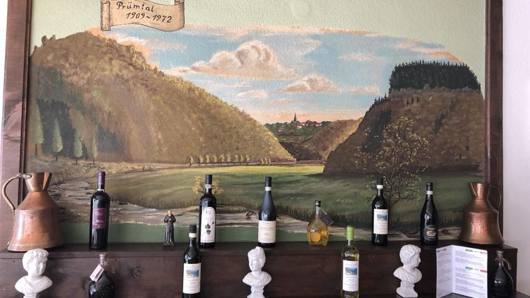 Een muurschildering met een landelijk landschap, omgeven door heuvels en een blauwe lucht. Daaronder staat een houten plank met verschillende wijnflessen en decoratieve elementen.