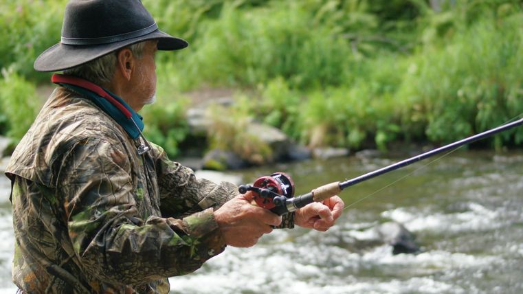 A man is fishing at the riverbank with a fishing rod. In the background, green plants and flowing water can be seen.