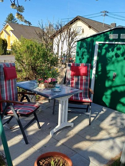 Ein gemütlicher Außenbereich mit einem Tisch und zwei Stühlen, umgeben von Pflanzen. Im Hintergrund steht ein grünes Gartenhaus und es scheint sonnig zu sein.