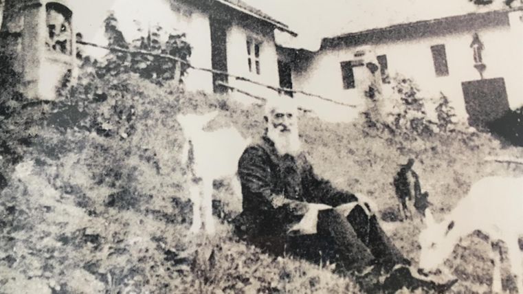 Ein älterer Mann sitzt auf einer Wiese neben Ziegen, mit einem Haus im Hintergrund. Die Szene wirkt ländlich und nostalgisch.