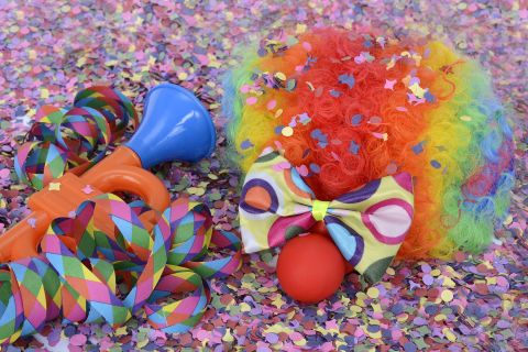 Ein bunter Clownhut mit vielen Farben und Glitzer. Neben ihm liegen eine bunte Trompete und Konfetti auf dem Boden.