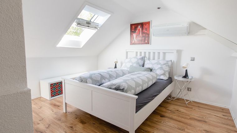 Ein modernes Schlafzimmer mit einem weißen Bett und gemütlicher Bettwäsche. Dazu gibt es ein Fenster und dekorative Wände in hellen Farben.