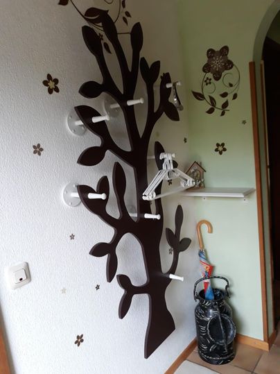 Un arbre à vêtements décoratif en forme d'arbre avec des crochets à vêtements blancs. En arrière-plan, des motifs floraux et un coin avec un porte-parapluie sont visibles.