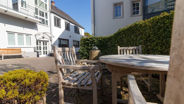 Ein gemütlicher Außenbereich mit Holzmöbeln und einer grünen Hecke. Die Sonne scheint über einem ruhigen Hof.