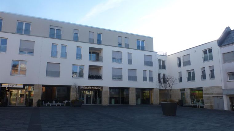 Ein modernes Gebäude mit weißen Fassaden und großen Fenstern. Der Platz ist gepflastert und bietet Platz für Sitzgelegenheiten im Freien.