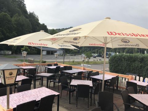 Een terras met tafels en stoelen, overdekt door grote parasols. Op de achtergrond zijn bomen en een rustige straat te zien.