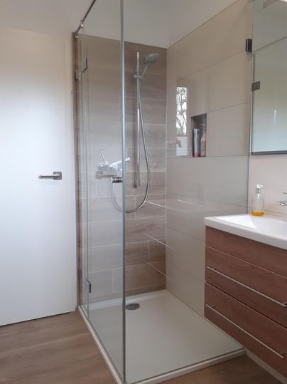 Salle de bain moderne avec une cabine de douche en verre et un lavabo avec meuble en bois. Des carreaux clairs et un miroir élégant créent une atmosphère accueillante.