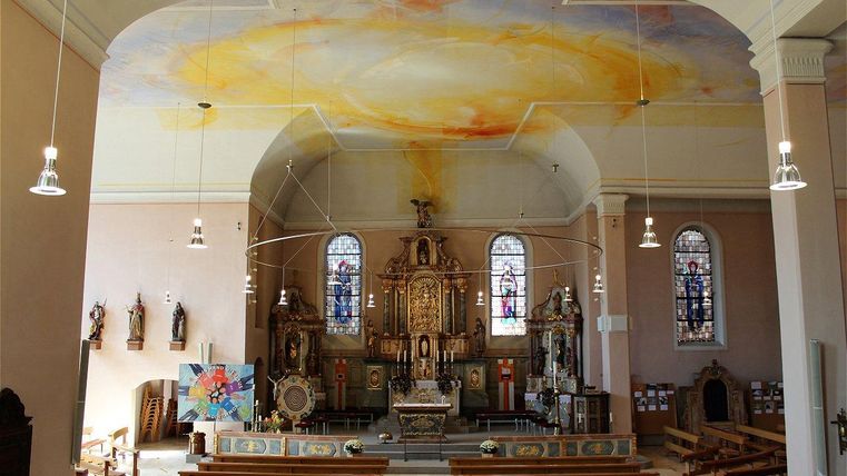 Une église sereine avec un impressionnant plafond peint et des vitraux colorés. Les bancs sont vides et l'autel se détache au centre de l'espace.
