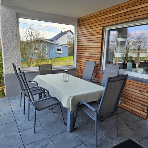 Une table à manger accueillante sur une terrasse couverte avec six chaises. À l'arrière-plan, on peut voir quelques arbres et une maison bleue.