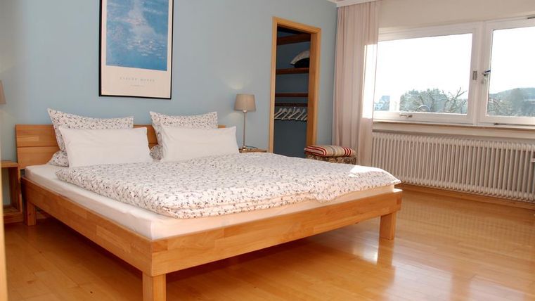 Ein helles Schlafzimmer mit einem großen Bett und komfortablen Kissen. Die Wände sind blau gestrichen und ein großes Fenster sorgt für viel Tageslicht.