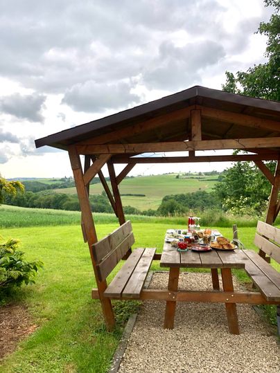 A cozy pavilion with a picnic table on a green meadow. In the background, gentle hills extend under a cloudy sky.