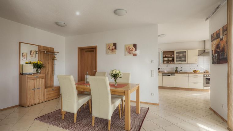 Ein heller Essbereich mit einem Holztablett und weißen Stühlen. Die offene Küche im Hintergrund ist modern und gut ausgestattet.