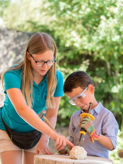 Een jongen en een meisje werken samen aan een blok hout. Beide dragen een veiligheidsbril en concentreren zich op hun taak.