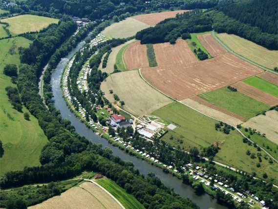 Eine Luftaufnahme von einer schlängelnden Flusslandschaft mit grünen Wäldern und Feldern. Kleine Campingplätze sind entlang des Ufers sichtbar.