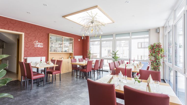 Ein modernes Restaurant mit rot-grauen Wänden und eleganten Tischen. Große Fenster sorgen für viel Tageslicht und eine einladende Atmosphäre.