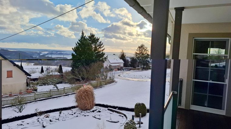 Un paysage d'hiver avec de la neige fraîchement tombée et un ciel dégagé. À l'arrière-plan, on peut voir des maisons et des arbres.