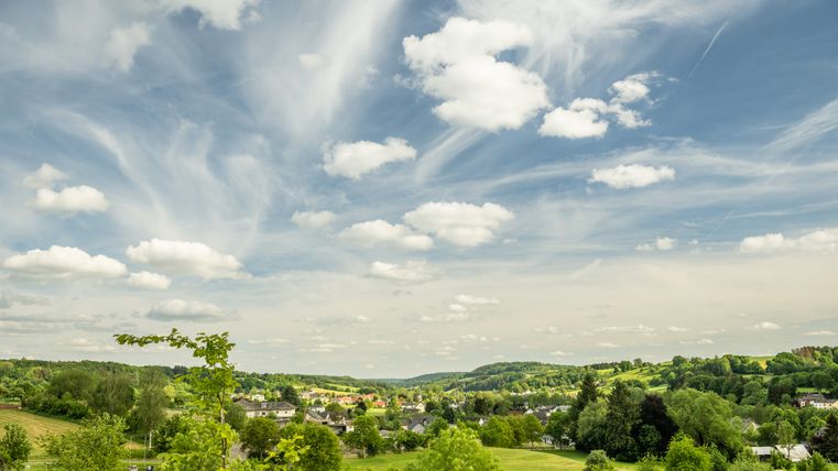 Landschaft mit grünen Feldern, Bäumen und einem Dorf unter einem blauen Himmel mit weißen Wolken.