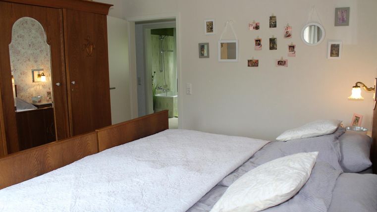 Ein gemütliches Schlafzimmer mit einem großen Bett und einer hölzernen Kommode. An der Wand hängen verschiedene Bilder, und ein Spiegel reflektiert das Licht.