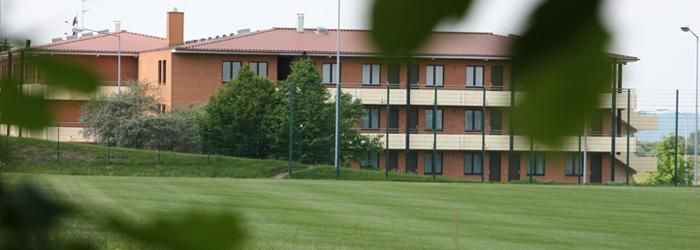 Een modern gebouw met meerdere verdiepingen staat in een groene omgeving. De lucht is helder en de omgeving is rustig.