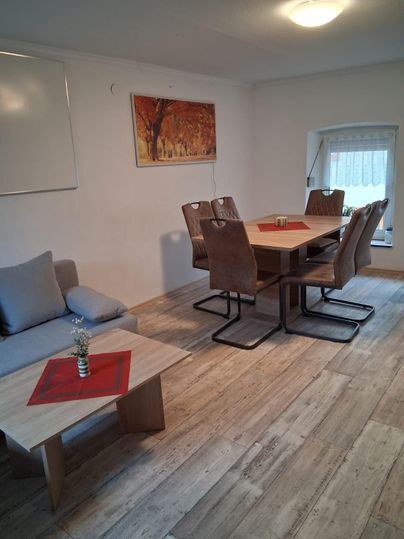 Une salle à manger moderne avec une grande table en bois et des chaises confortables. Un tableau coloré est accroché au mur, et une petite table avec une fleur est placée au premier plan.