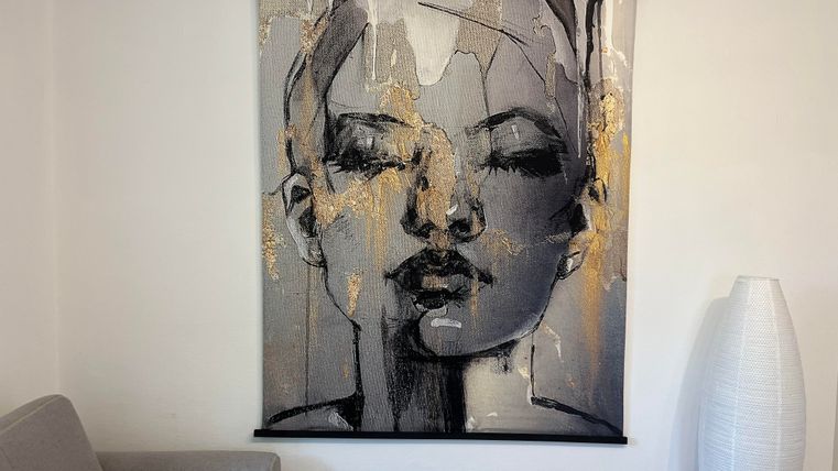 Une œuvre murale moderne avec un portrait abstrait d'une femme dans des tons noir et blanc et des accents dorés. Elle est accrochée à un mur à côté d'un canapé gris et d'une lampe sur pied blanche.