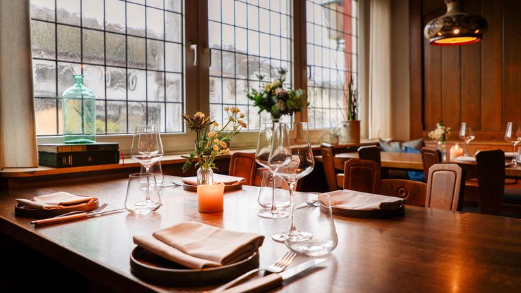 Un restaurant confortable avec une table en bois, des verres élégants et des fleurs sur la table. Les grandes fenêtres laissent entrer une lumière douce et créent une atmosphère accueillante.