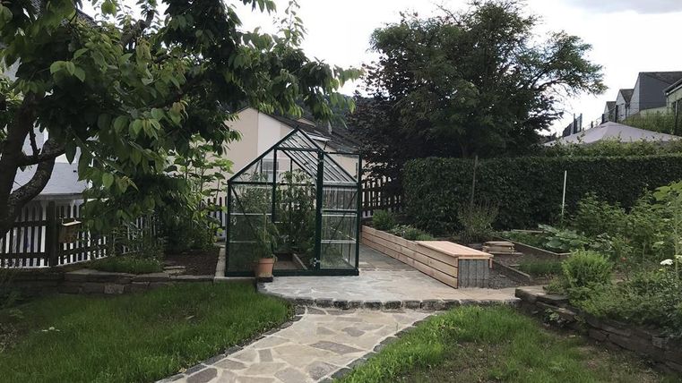 Ein schöner Garten mit einem Gewächshaus und einer gepflasterten Fläche. Umgeben von Bäumen und einer Hecke, bietet er eine grüne Oase.