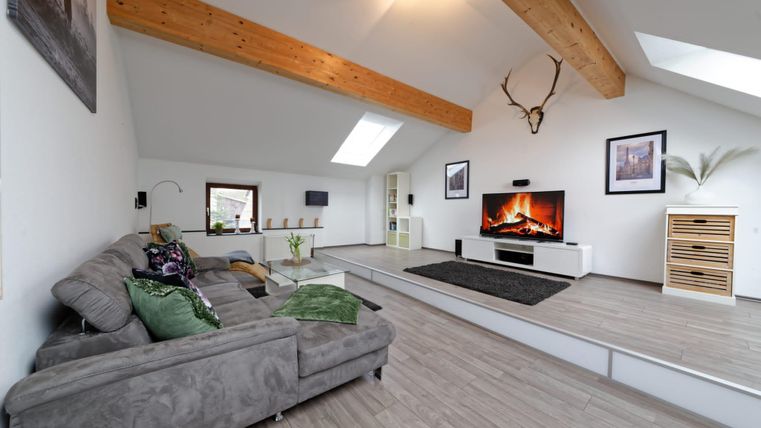 Un salon moderne avec un canapé gris et un grand téléviseur à écran plat. De grandes fenêtres et des poutres en bois donnent à la pièce une atmosphère lumineuse et accueillante.