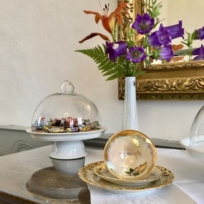 Une belle table avec une cloche en verre pleine de bonbons et une élégante tasse en porcelaine. À l'arrière-plan, il y a des fleurs fraîches dans un vase blanc et un cadre doré au mur.
