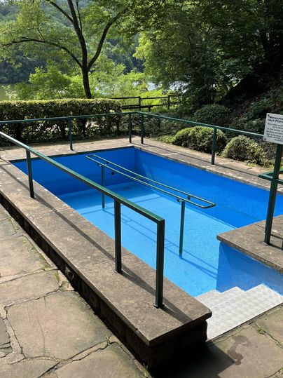 Ein kleiner, blauer Pool mit Geländern in einer grünen Umgebung. Im Hintergrund sind Bäume und ein ruhiger See sichtbar.