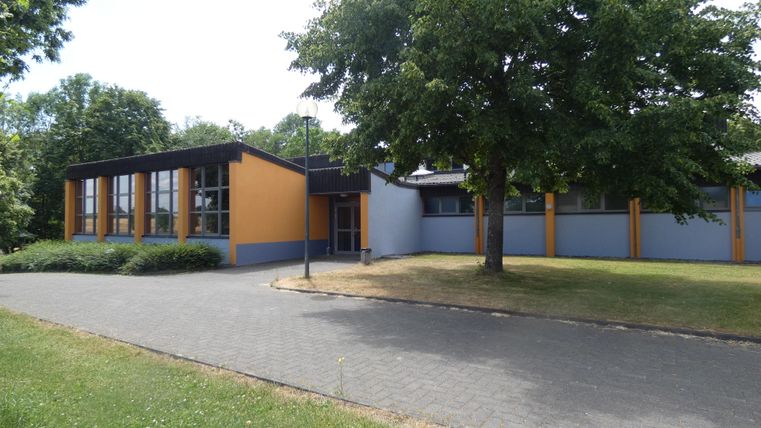 Ein modernes Gebäude mit orangefarbenen und grauen Wänden, umgeben von Bäumen und einem gepflegten Rasen. Der Zugang erfolgt über einen gepflasterten Weg.
