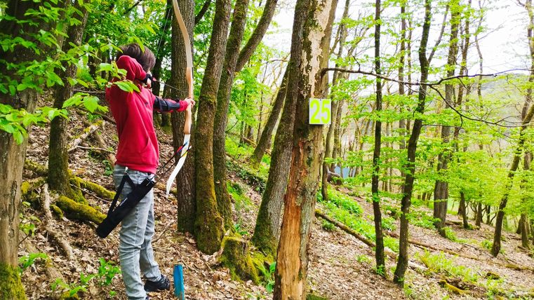 Een persoon schiet met een boog in het bos. De omgeving is groen en nodigt uit tot actieve recreatie.