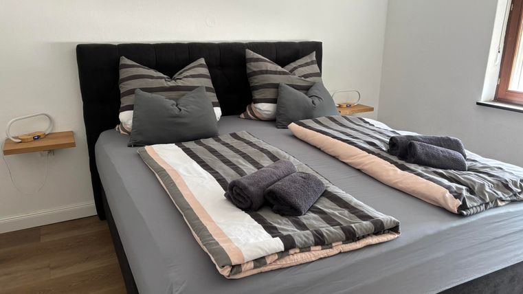 Ein modernes Schlafzimmer mit einem großen Doppelbett und gestreiften Kissen. Die Bettwäsche ist in Grau und Beige gehalten, und es gibt zwei Handtücher auf dem Bett.