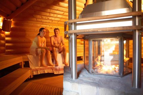 Eine gemütliche Sauna mit warmem Licht und Holzverkleidung. Zwei Personen sitzen entspannt auf den Bänken neben einem Kamin.