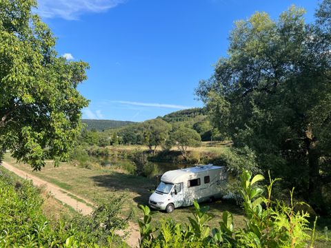 Un camping-car est stationné dans un paysage calme et verdoyant. À l'arrière-plan, on voit des arbres et un ciel bleu.